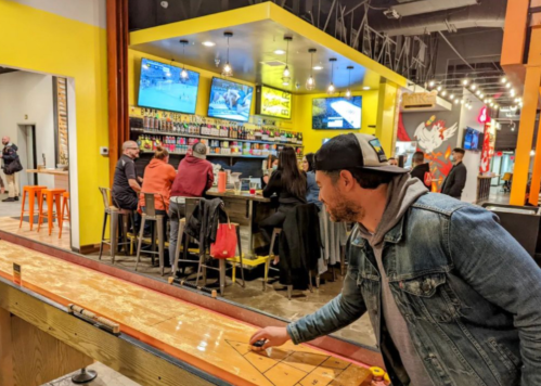 A man plays shuffleboard in a vibrant bar with multiple screens and patrons seated at the counter.